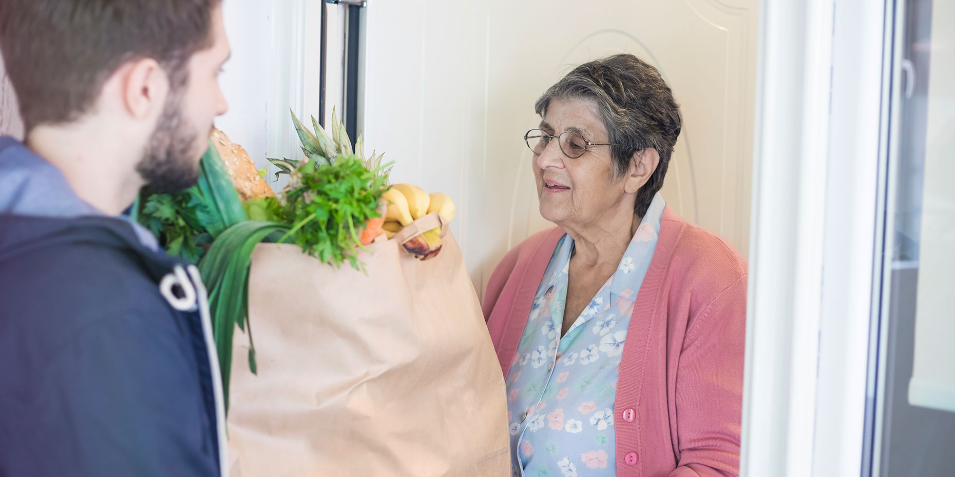 Senior Woman Answers the door smiling as a young man hands her a grocery bag full of fresh produce.