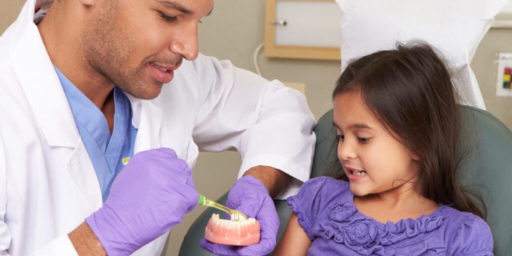 Pediatric dentist teaching young child how to brush teeth with a model of teeth