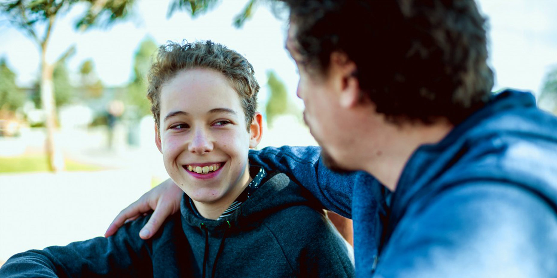 Young man sitting smiling looking at person out of focus looking at him with his arm around him