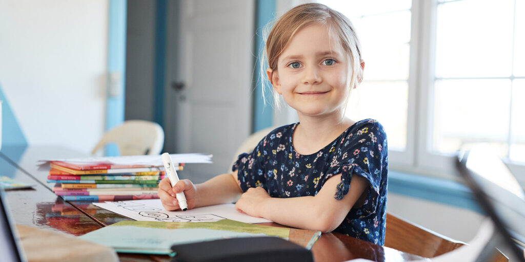 Child looking directly into the camera and smiling while coloring at her desk with a marker